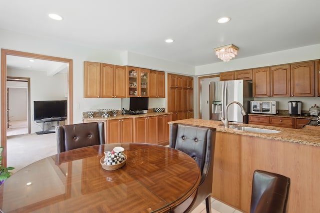 kitchen with a sink, stainless steel fridge with ice dispenser, glass insert cabinets, light stone counters, and brown cabinets