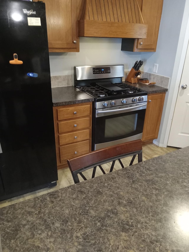 kitchen featuring gas range, freestanding refrigerator, premium range hood, dark countertops, and brown cabinets