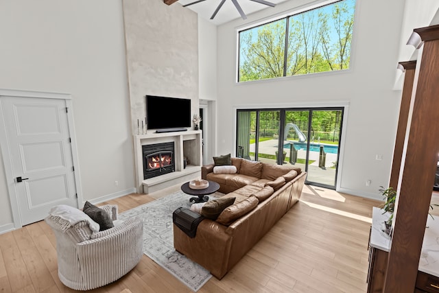 living room with a high ceiling, a glass covered fireplace, ceiling fan, light wood finished floors, and baseboards