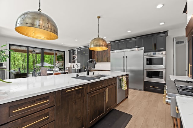 kitchen with appliances with stainless steel finishes, a sink, light wood finished floors, hanging light fixtures, and light stone counters