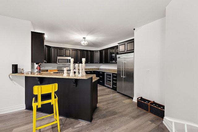kitchen featuring appliances with stainless steel finishes, wood finished floors, a peninsula, a kitchen bar, and light stone countertops