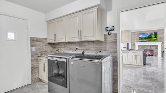 clothes washing area featuring tile walls, cabinet space, washing machine and dryer, light marble finish floors, and a glass covered fireplace