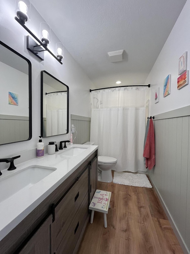 bathroom with wood finished floors, wainscoting, toilet, a textured ceiling, and double vanity