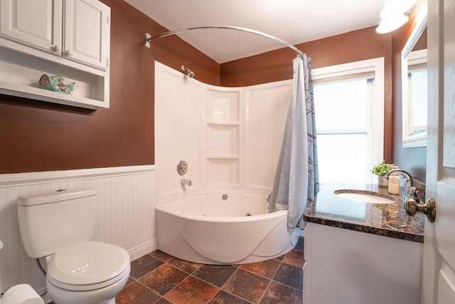 bathroom with stone tile flooring, shower / bathtub combination with curtain, a wainscoted wall, toilet, and vanity