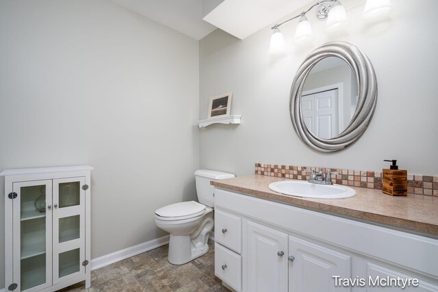 half bathroom featuring toilet, vanity, baseboards, tasteful backsplash, and stone finish flooring