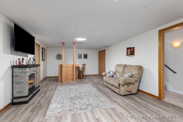 living area with light wood-type flooring, baseboards, a drop ceiling, and a stone fireplace