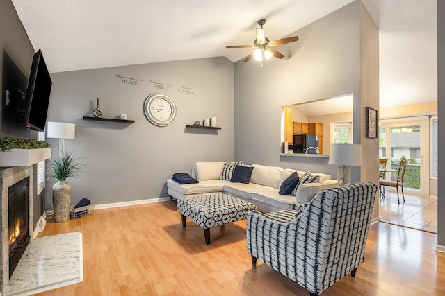 living room with vaulted ceiling, a tiled fireplace, ceiling fan, light wood finished floors, and baseboards