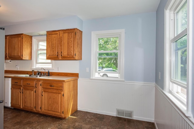 kitchen with a sink, healthy amount of natural light, wainscoting, brown cabinetry, and light countertops