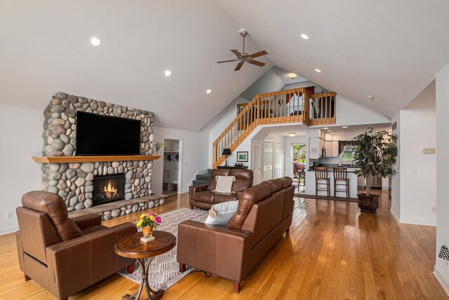 living room featuring a ceiling fan, high vaulted ceiling, light wood-style floors, stairway, and a stone fireplace