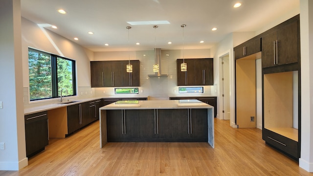 kitchen with wall chimney range hood, a sink, dark brown cabinets, a kitchen island, and light wood-style floors