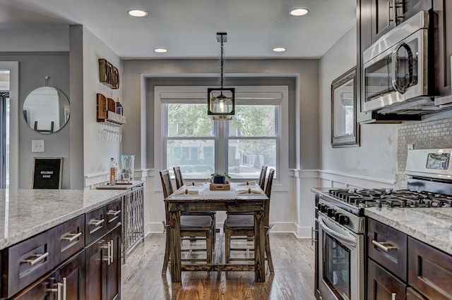 kitchen with stainless steel appliances, dark brown cabinets, light stone counters, light wood-style floors, and a wainscoted wall