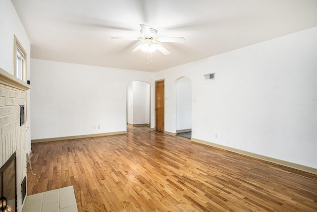 unfurnished living room with a brick fireplace, arched walkways, light wood-style floors, and ceiling fan