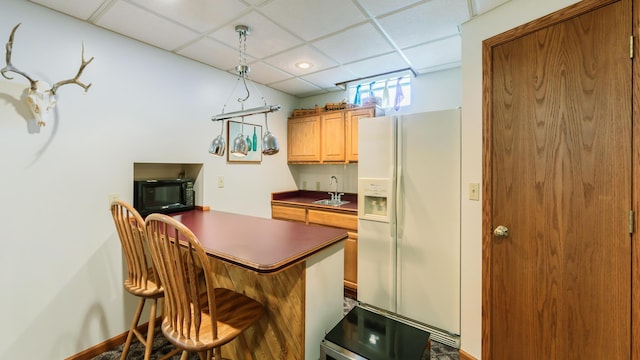 kitchen with white refrigerator with ice dispenser, black microwave, dark countertops, a paneled ceiling, and a sink