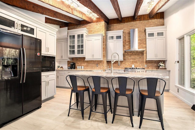kitchen with black appliances, wall chimney range hood, a sink, beamed ceiling, and light wood-type flooring