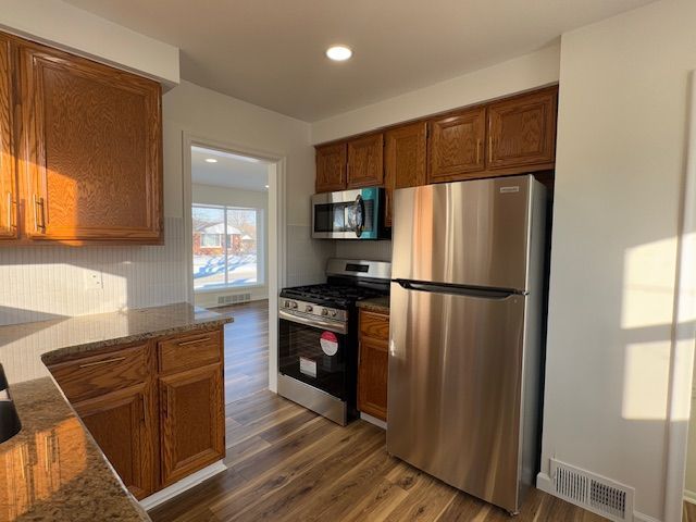 kitchen with appliances with stainless steel finishes, dark wood-type flooring, brown cabinets, stone countertops, and recessed lighting