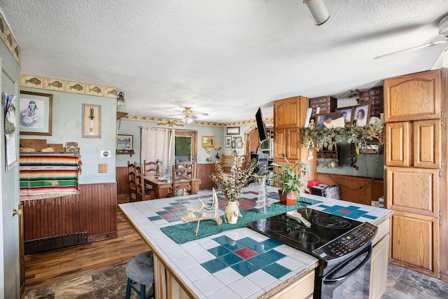 kitchen with tile counters, ceiling fan, a textured ceiling, wainscoting, and wooden walls