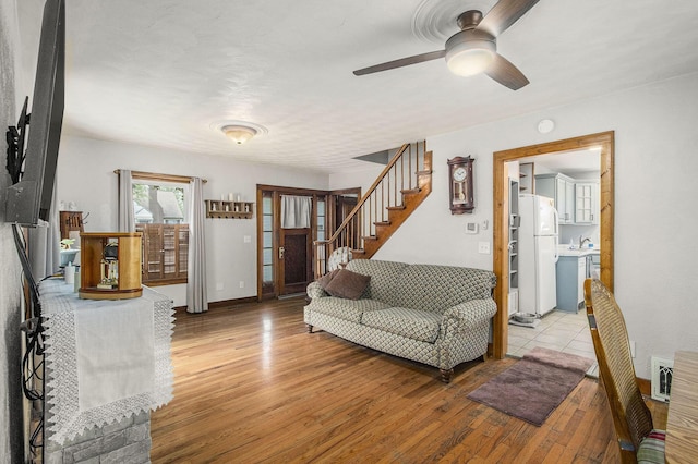 living area with light wood-type flooring, ceiling fan, stairway, and baseboards