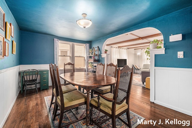 dining room featuring wainscoting, wood finished floors, arched walkways, and beamed ceiling
