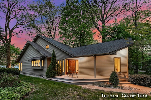 back of house at dusk with a wooden deck, roof with shingles, and a lawn