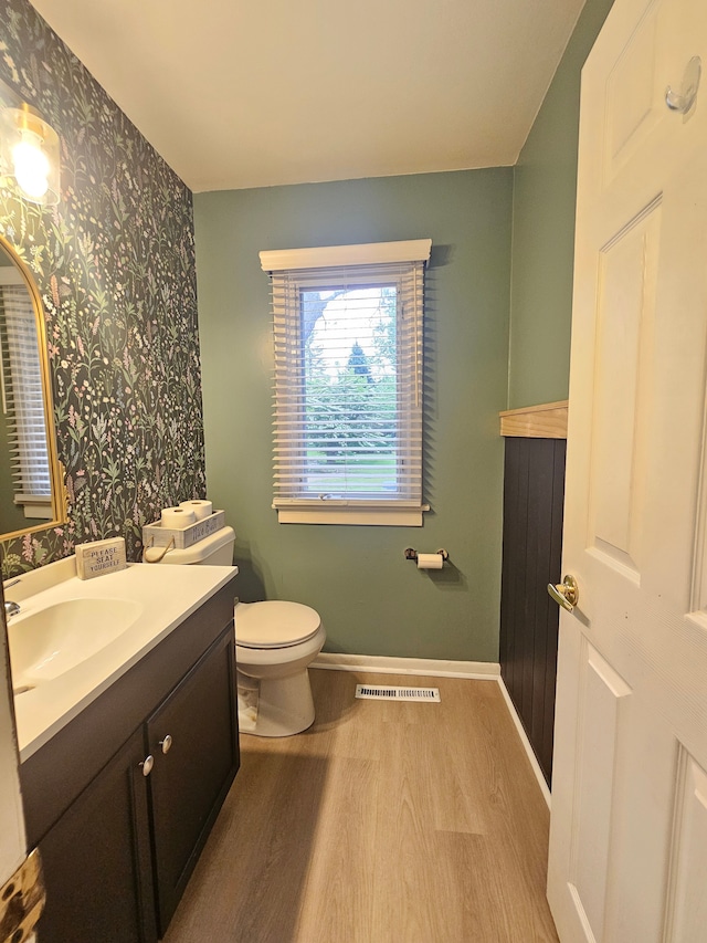 half bath with wallpapered walls, wood finished floors, vanity, toilet, and baseboards