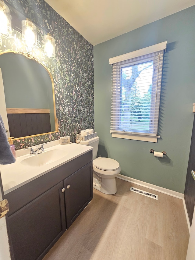 bathroom with wallpapered walls, wood finished floors, toilet, baseboards, and vanity