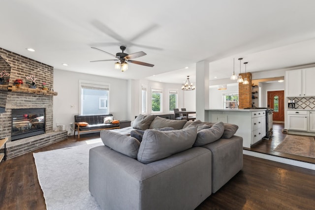 living room featuring a brick fireplace, dark wood-style floors, a ceiling fan, recessed lighting, and a chandelier