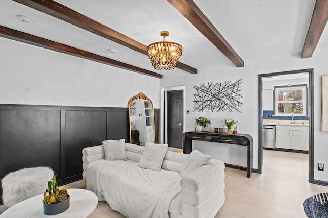 living room featuring a chandelier, beamed ceiling, light wood-style floors, and a wainscoted wall