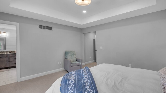 bedroom with a raised ceiling, light colored carpet, baseboards, and connected bathroom