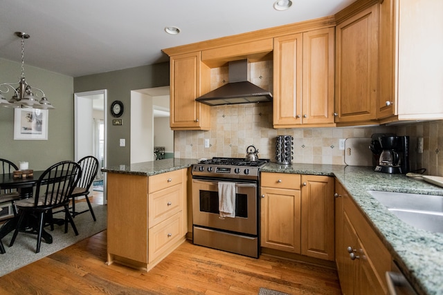 kitchen featuring stainless steel appliances, wall chimney range hood, a chandelier, light wood-style flooring, and dark stone counters