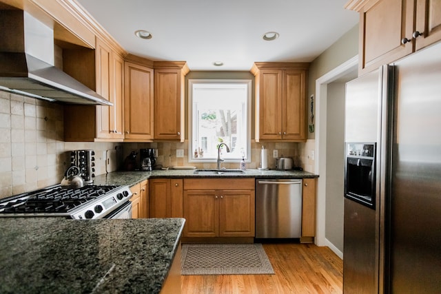 kitchen featuring appliances with stainless steel finishes, a sink, wall chimney exhaust hood, light wood-style floors, and dark stone counters