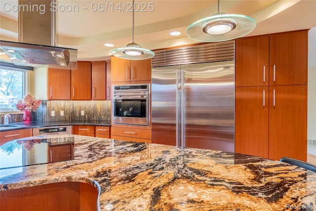12950 Via Catherina Dr, Grand Blanc, MI 48439 - kitchen featuring stainless steel appliances, stone countertops, brown cabinets, range hood, and decorative backsplash