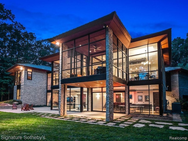 6956 Cooley Lake Rd, White Lake, MI 48383 - back of house at night featuring a patio, metal roof, stone siding, a lawn, and a sunroom