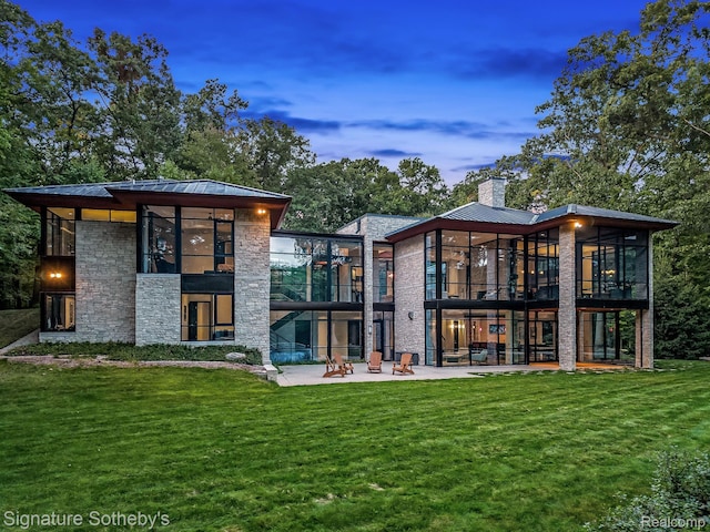 6956 Cooley Lake Rd, White Lake, MI 48383 - back of house at dusk featuring stone siding, a patio, a sunroom, a chimney, and a yard
