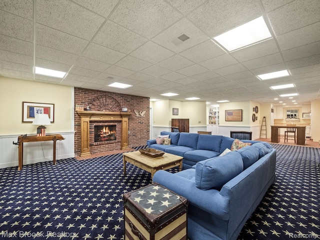 carpeted living room featuring a fireplace, a drop ceiling, and a wainscoted wall