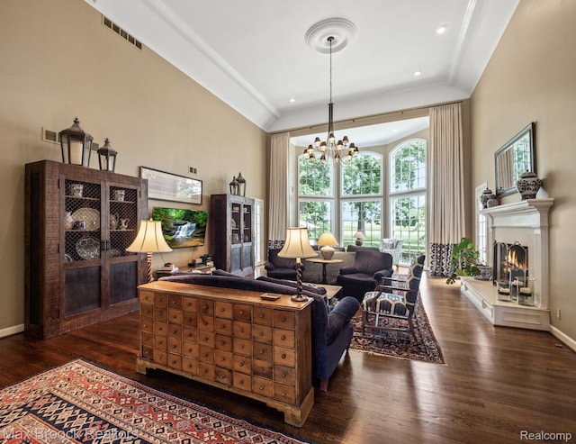 living area with dark wood-style flooring, a towering ceiling, a chandelier, a warm lit fireplace, and crown molding
