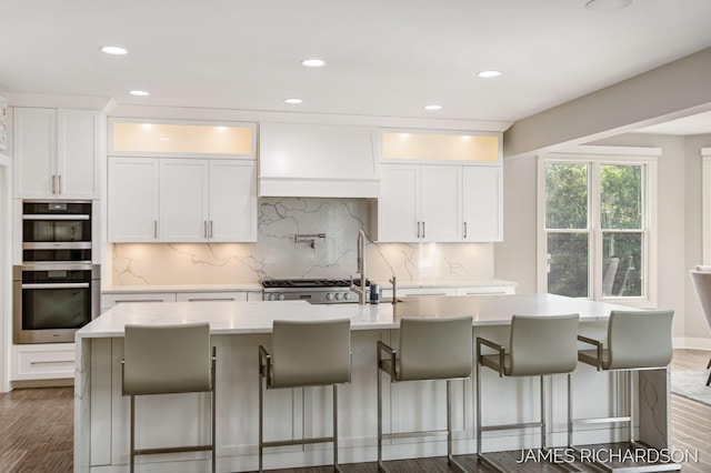 12974 Boulderway Trl, Grand Haven, MI 49417 - kitchen with double wall oven, custom range hood, a center island with sink, white cabinets, and dark wood-style floors
