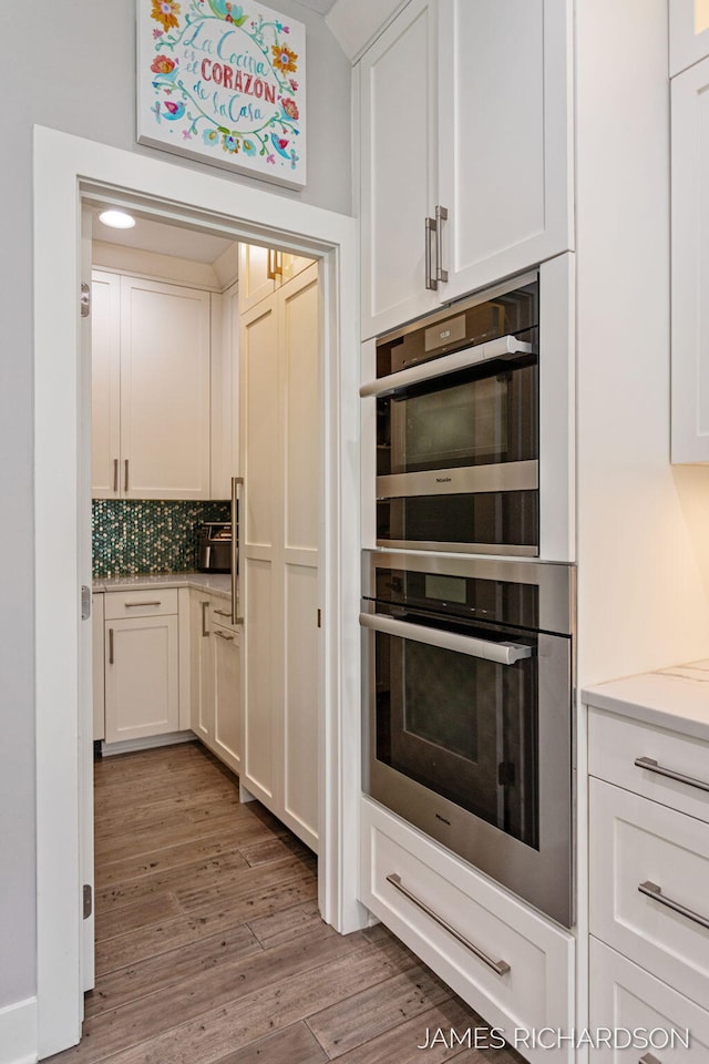 12974 Boulderway Trl, Grand Haven, MI 49417 - kitchen featuring stainless steel double oven, white cabinetry, wood finished floors, decorative backsplash, and light countertops