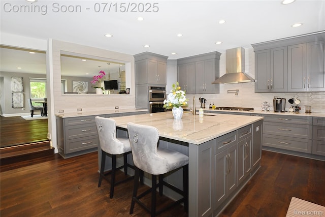 591 Wallace St, Birmingham, MI 48009 - kitchen featuring wall chimney exhaust hood, gray cabinetry, an island with sink, decorative backsplash, and a breakfast bar area
