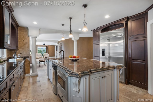 56 Oxford Rd, Grosse Pointe Shores, MI 48236 - kitchen featuring decorative columns, built in appliances, an island with sink, dark stone counters, and dark brown cabinetry