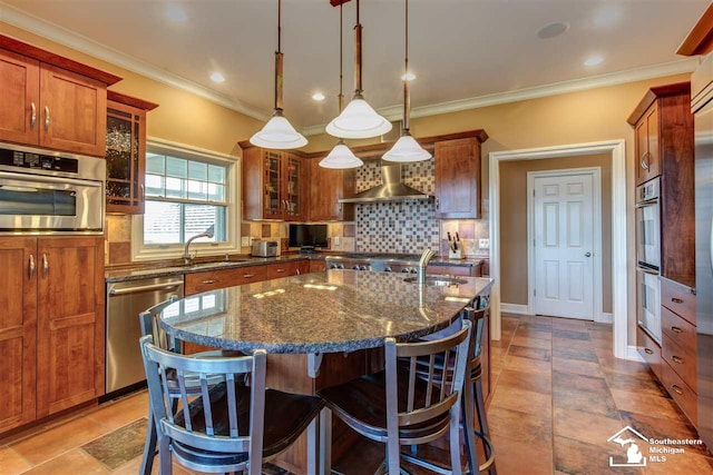 kitchen featuring wall chimney exhaust hood, hanging light fixtures, backsplash, an island with sink, and appliances with stainless steel finishes