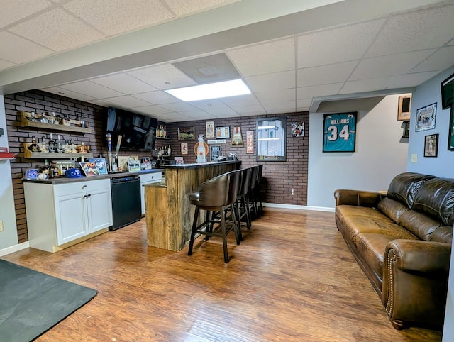 bar featuring black dishwasher, a dry bar, a paneled ceiling, brick wall, and wood finished floors