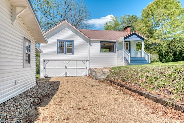 view of front of property with dirt driveway, covered porch, a chimney, metal roof, and an attached garage