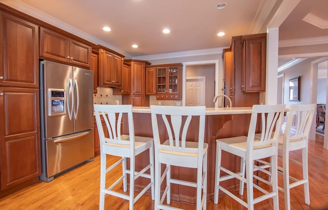 kitchen with crown molding, stainless steel refrigerator with ice dispenser, light wood finished floors, brown cabinetry, and a kitchen breakfast bar