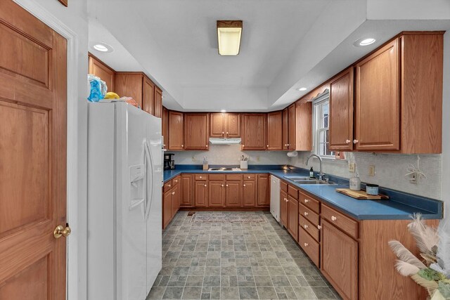 kitchen featuring dark countertops, backsplash, brown cabinetry, white appliances, and a sink