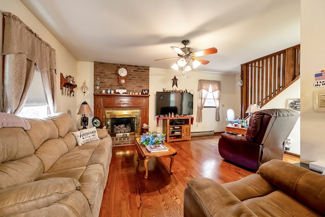 living room featuring stairway, a fireplace, wood finished floors, baseboard heating, and ceiling fan