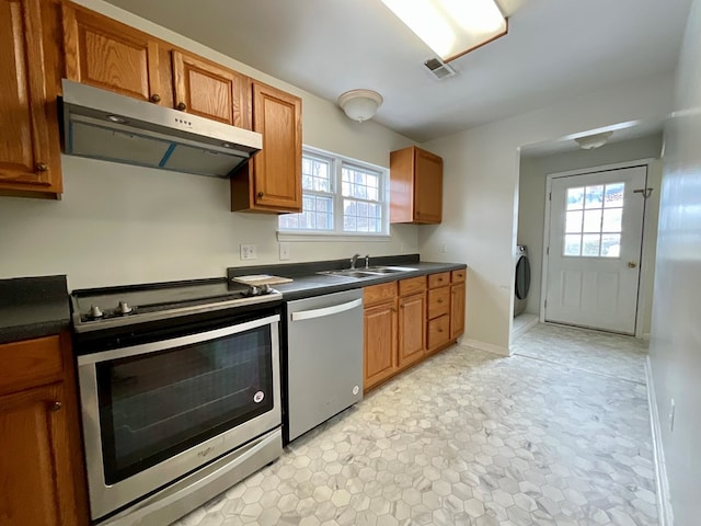 kitchen with washer / clothes dryer, stainless steel appliances, dark countertops, under cabinet range hood, and brown cabinetry
