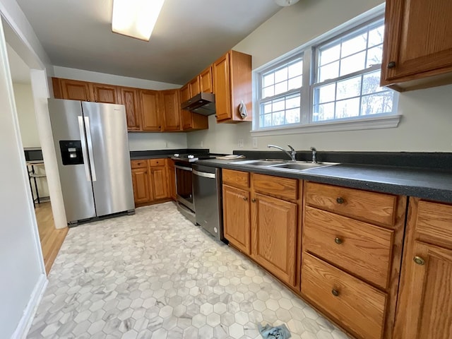 kitchen with a sink, appliances with stainless steel finishes, brown cabinetry, under cabinet range hood, and dark countertops