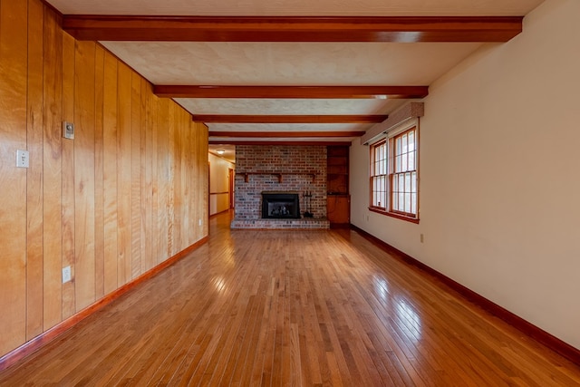 unfurnished living room with a brick fireplace, baseboards, wood walls, beam ceiling, and hardwood / wood-style flooring