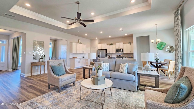 living room with ceiling fan with notable chandelier, a raised ceiling, light wood-type flooring, and crown molding