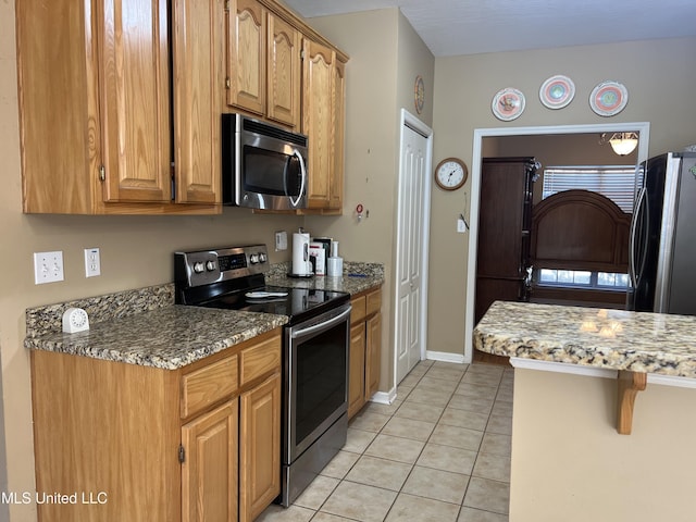 kitchen with light tile patterned floors, brown cabinetry, baseboards, stone counters, and appliances with stainless steel finishes